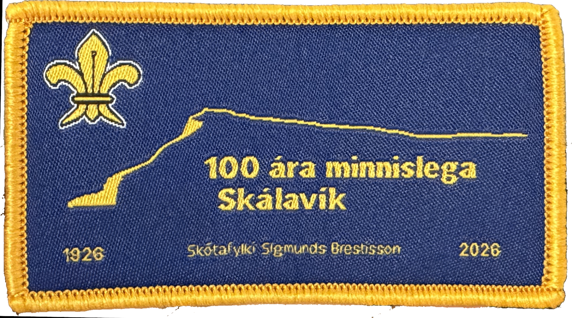 Vævet rektangulært mærke '100 ára minnislega Skálavík 1926–2026' med gul kant og spejderlilje